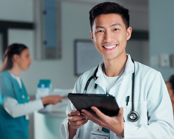 A smiling doctor in a white coat is holding a tablet in their hand. In the background, two other medical professionals can be seen talking to each other.