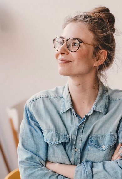 A smiling woman with glasses sits in a bright room, wearing a denim shirt and with her arms crossed. She looks thoughtful and appears content.