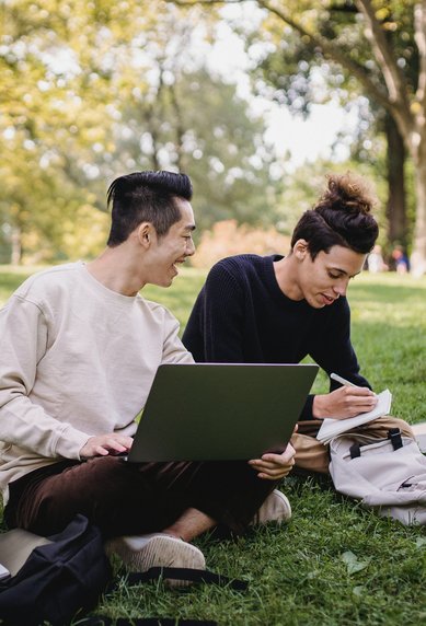 Zwei Personen sitzen auf einer Wiese, einer arbeitet am Laptop, während der andere Notizen macht. Um sie herum sind Bäume und eine entspannte Atmosphäre.
