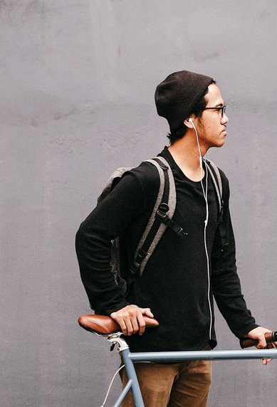 A young man stands next to a bicycle in front of a gray wall. He is wearing a black sweater, a beanie, and has a backpack on his back.
