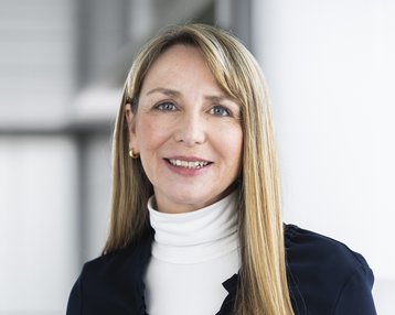 A woman with long, straight hair is wearing a dark blue top and a white turtleneck sweater. She is smiling warmly.