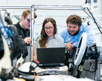 Drei Personen betrachten gemeinsam einen Laptop, während sie an einem technischen Projekt in einer modernen Werkstatt arbeiten.