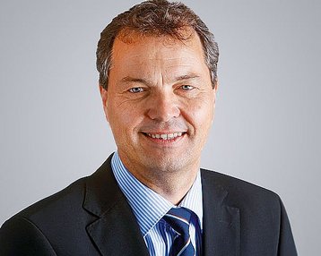 A man with short brown hair and a friendly smile is wearing a black suit and a striped shirt. The background is neutral and bright.