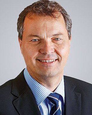 A man with short brown hair and a friendly smile is wearing a black suit and a striped shirt. The background is neutral and bright.
