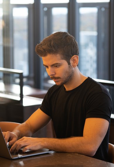 Ein junger Mann sitzt an einem Tisch und arbeitet konzentriert an einem Laptop in einem modernen, hellen Raum mit großen Fenstern.