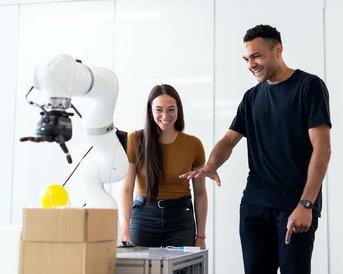 Zwei Personen stehen an einem Tisch und beobachten einen Roboterarm, der eine gelbe Kugel greift. Beide wirken interessiert und erfreut.