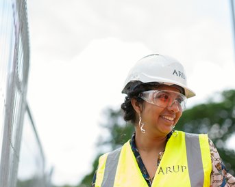 Eine lächelnde Frau in einem Sicherheitshelm und einer Warnweste steht an einer Baustelle, umgeben von einem Bauzaun und Bäumen im Hintergrund.