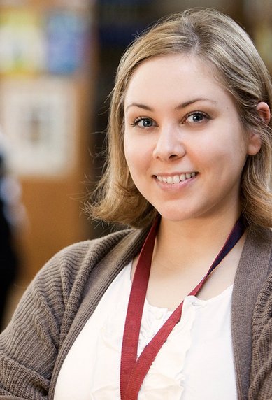 A young woman stands in a library with her arms crossed, smiling. In the background, other people can be seen working at tables.