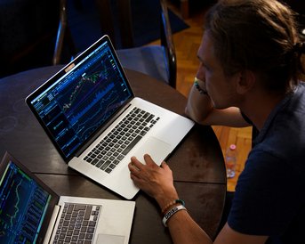 Ein Mann sitzt an einem Tisch mit zwei Laptops, die Finanzdiagramme anzeigen. Er schaut konzentriert auf die Bildschirme.