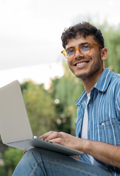 A young man with glasses is sitting outside, smiling as he types on a laptop. In the background, trees and a building can be seen.