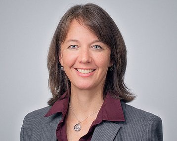 A woman with shoulder-length brown hair is wearing a gray blazer and a purple shirt. She is smiling warmly at the camera.
