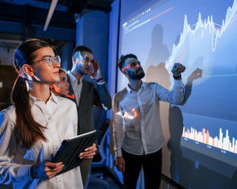 A group of professionals is looking at graphics and data on a large screen. A woman is holding a tablet and appears focused, while the others are discussing and pointing at the presentation.